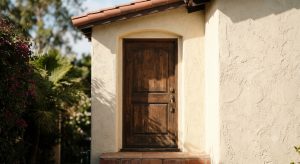 Close-up of a stucco home's wood front door and brass deadbolt in summer shade, illustrating an Orange County escrow, title, and closing costs guide.