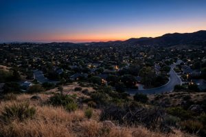 North Orange County foothill neighborhood at blue hour in midsummer, viewed from an elevated vantage with warm window light across the rooftops.
